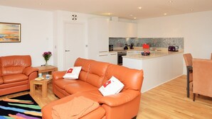 A modern living room with a sofa and a coffee table, a small kitchen area with a tiled backsplash, and a dining area with a table and chairs. - LUXURY APARTMENT PORTH, SLEEPS 4, SEA VIEWS OVERLOOKING WHIPSDERRY BEACH (Porth, Newquay)