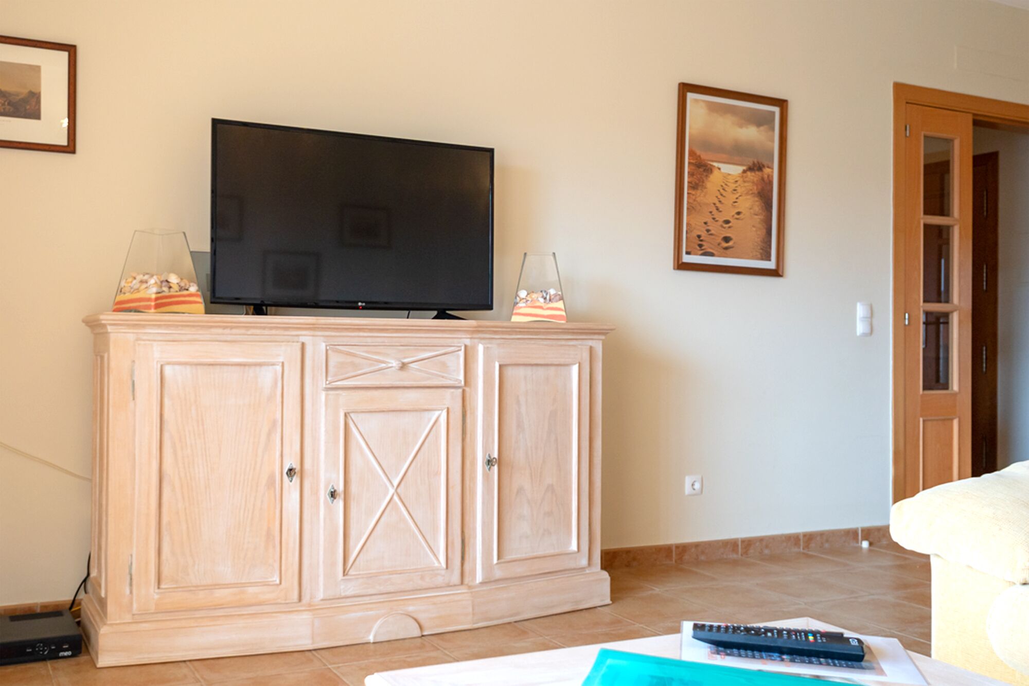 Ein Wohnzimmer mit Fernseher auf einer Holzkommode, einer Fernbedienung auf einer Zeitschrift und einer Tür im Hintergrund.