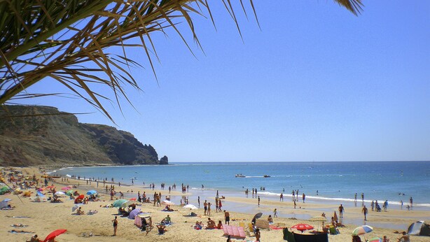 Una playa concurrida donde la gente se toma el sol y nada, un bote en el agua y un acantilado al fondo.