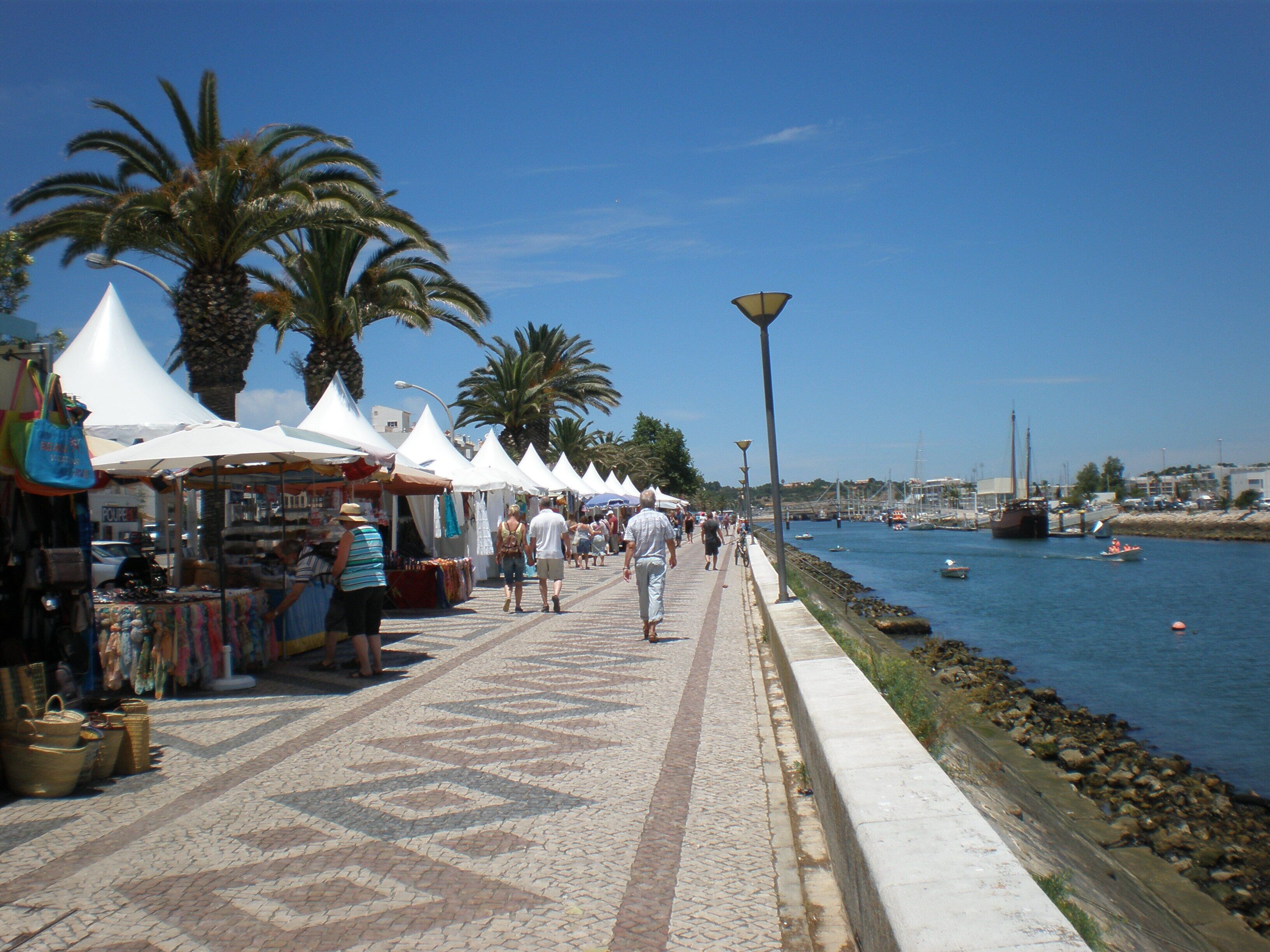 Une promenade en bord de mer bordée de boutiques et de palmiers, une allée dallée et un port tranquille où sont amarrés des bateaux.