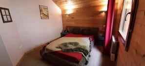 Een gezellige kamer met een houten plafond, een bed met een rode deken, een rieten stoel en een kleine tafel met een lamp.