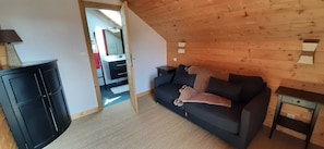 Une pièce chaleureuse avec un canapé sombre, une petite table et un meuble. On peut voir un plafond en bois et une porte qui mène à une autre pièce.