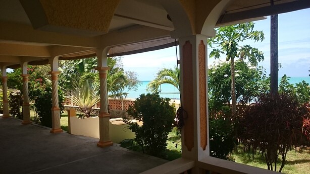 A covered outdoor area with a view of the sea, palm trees, and a garden.