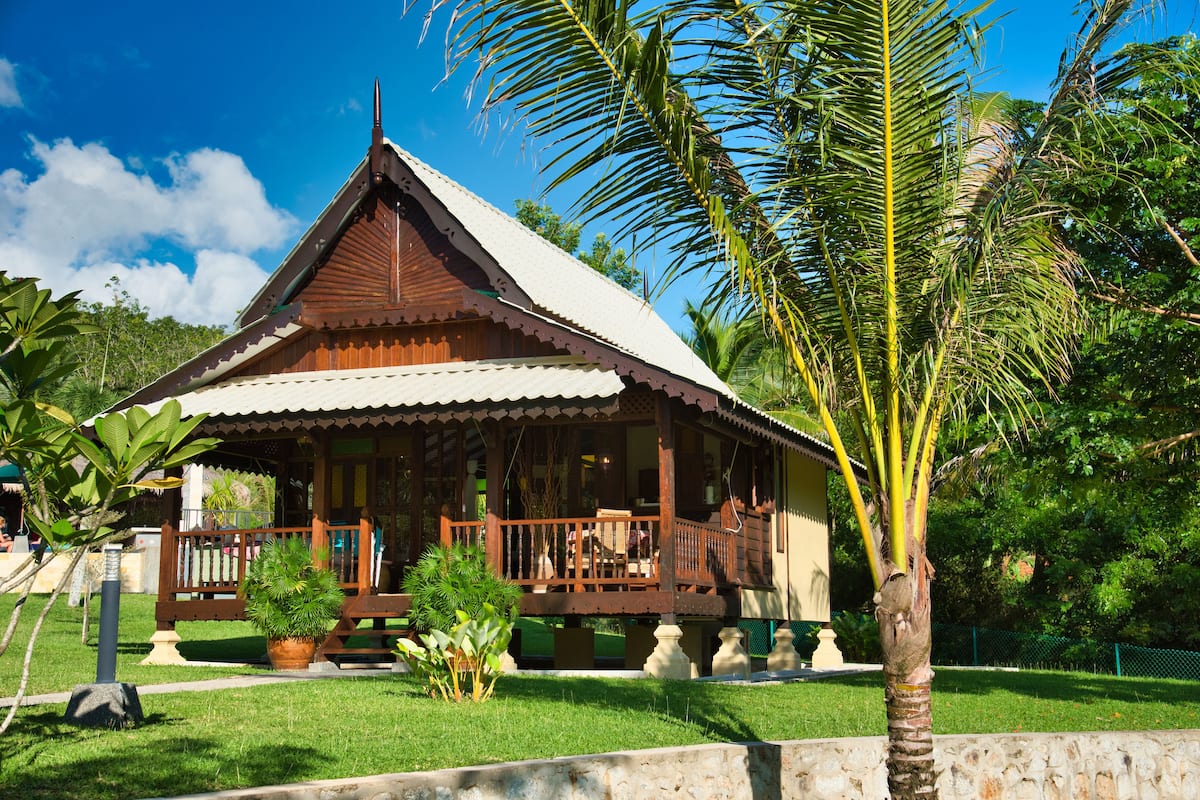 A traditional wooden house with a balcony, surrounded by lush greenery and palm trees.