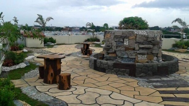 En stenbelagt terrasse med en central stenskulptur, omgivet af en anlagt have med forskellige planter og en stensti.