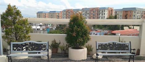 A rooftop terrace with two benches, potted plants, and a view of residential buildings.