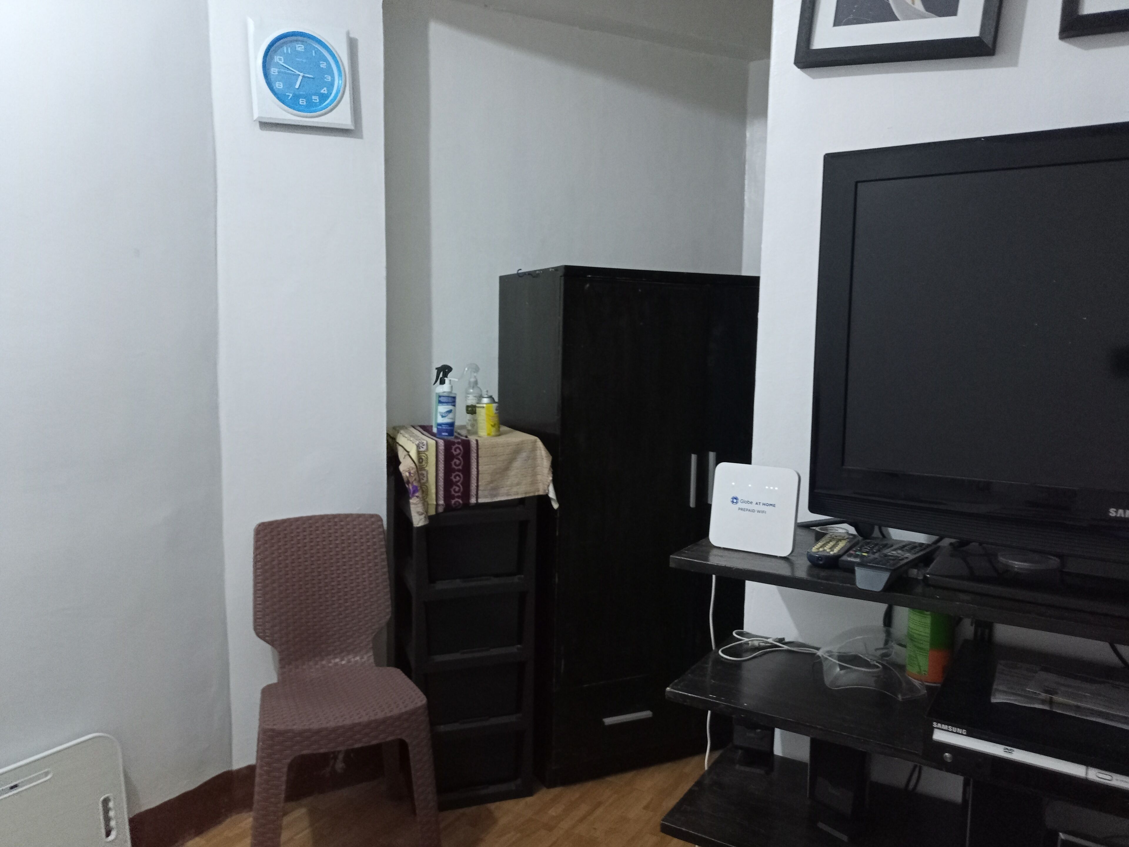 A small room with a black TV on a stand, a chair, a small table with cleaning supplies, and a clock on the wall.