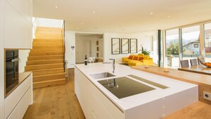 Une cuisine moderne avec un îlot central, un escalier en bois et un canapé jaune vif dans un espace de vie spacieux doté de grandes fenêtres.