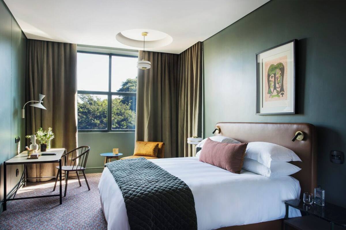 Une chambre d’hôtel avec un grand lit, un bureau avec une chaise, une petite table et une fenêtre avec des rideaux.
