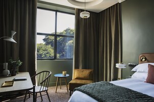 Habitación de hotel con cama, escritorio con silla, una mesita, lámpara y vistas a árboles por la ventana.