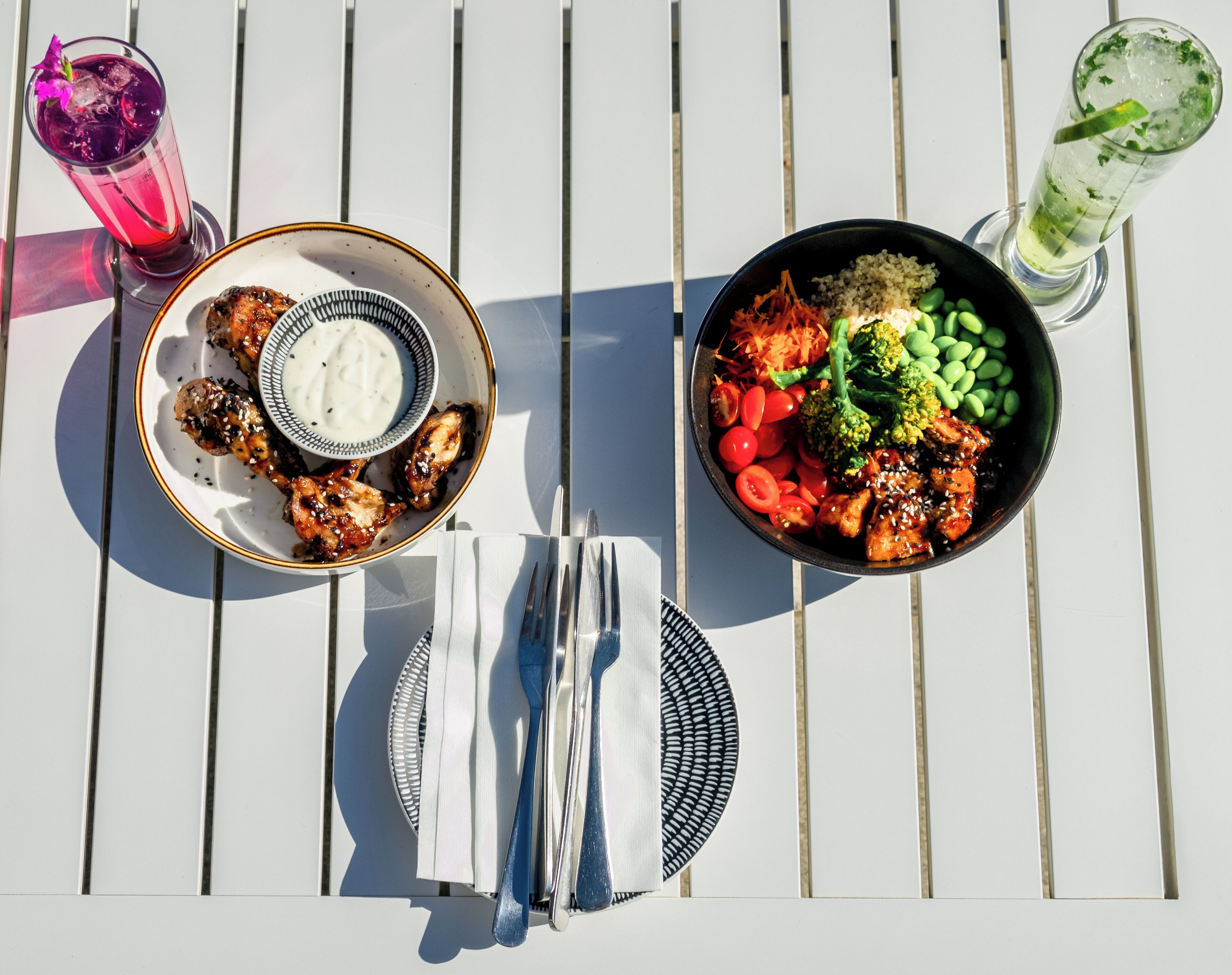 Una mesa con comida que incluye alitas de pollo a la parrilla, un tazón de arroz con vegetales y una bebida.