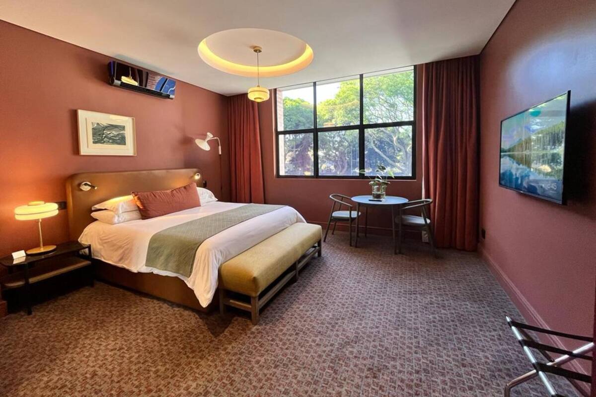 Une chambre d’hôtel comprenant un lit, une table de chevet, une lampe, une télévision, une fenêtre et une petite table avec des chaises.