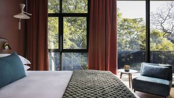 A hotel room with a large bed, a side table, a chair, and a view of trees through large windows.