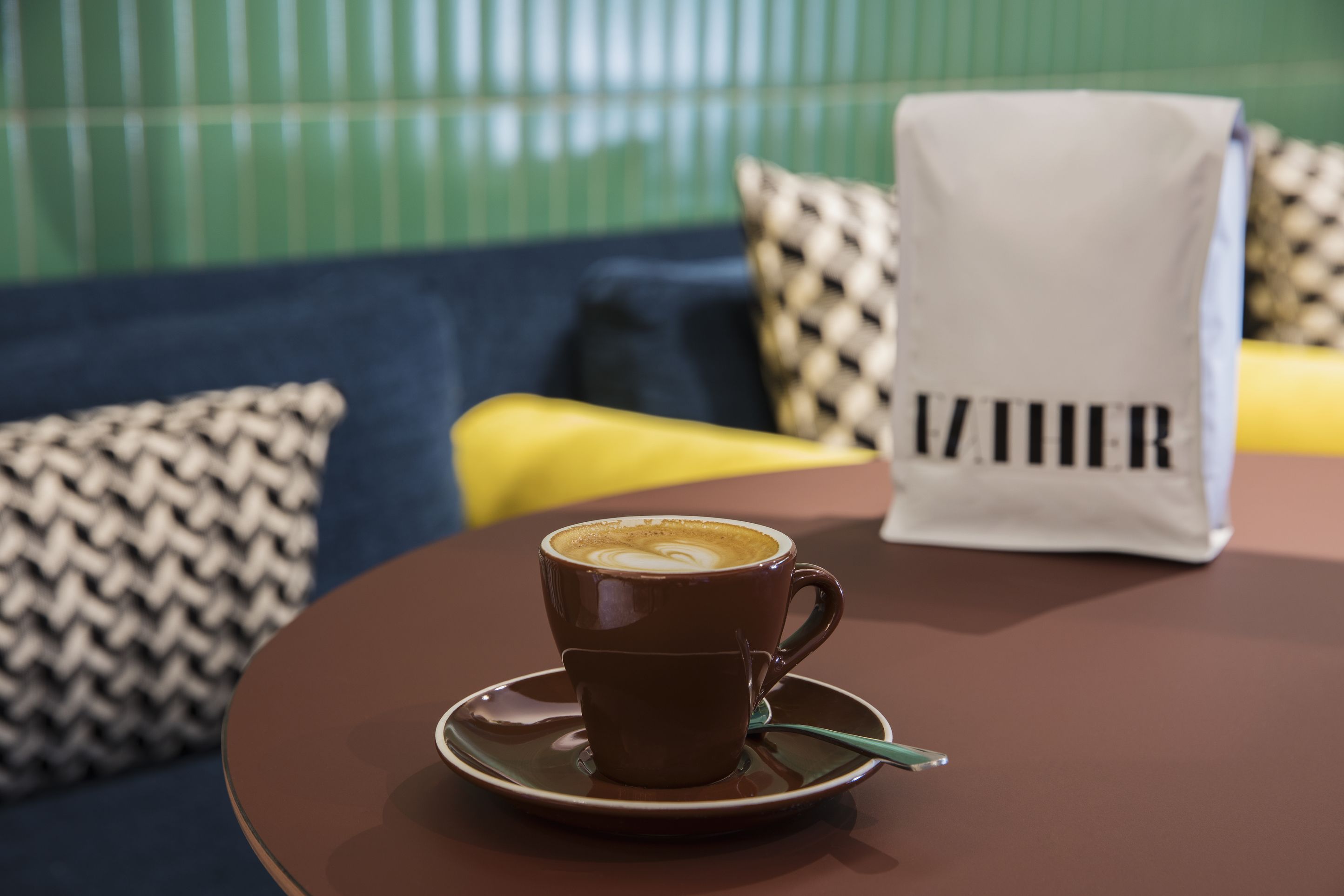 Uma chávena de café sobre um pires numa mesa, com uma mala com a etiqueta 'ZURICH' ao fundo.