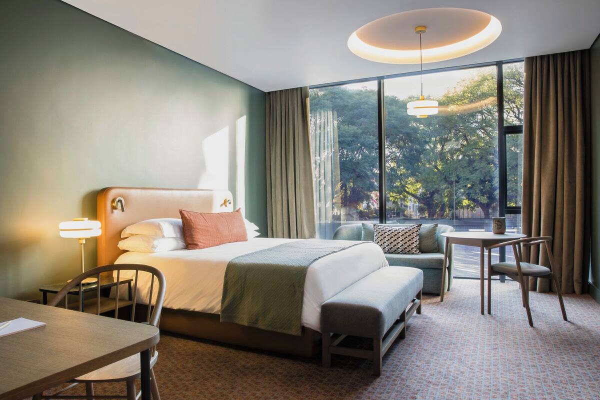 Une chambre d’hôtel avec un grand lit, un bureau, une chaise et une vue sur les arbres par de grandes fenêtres.