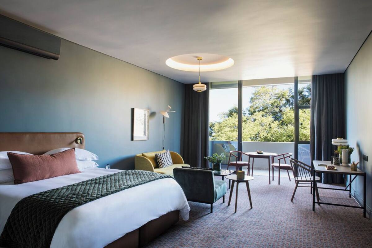 Une chambre d’hôtel avec un grand lit, un canapé, une table à manger avec des chaises et un balcon donnant sur la verdure.