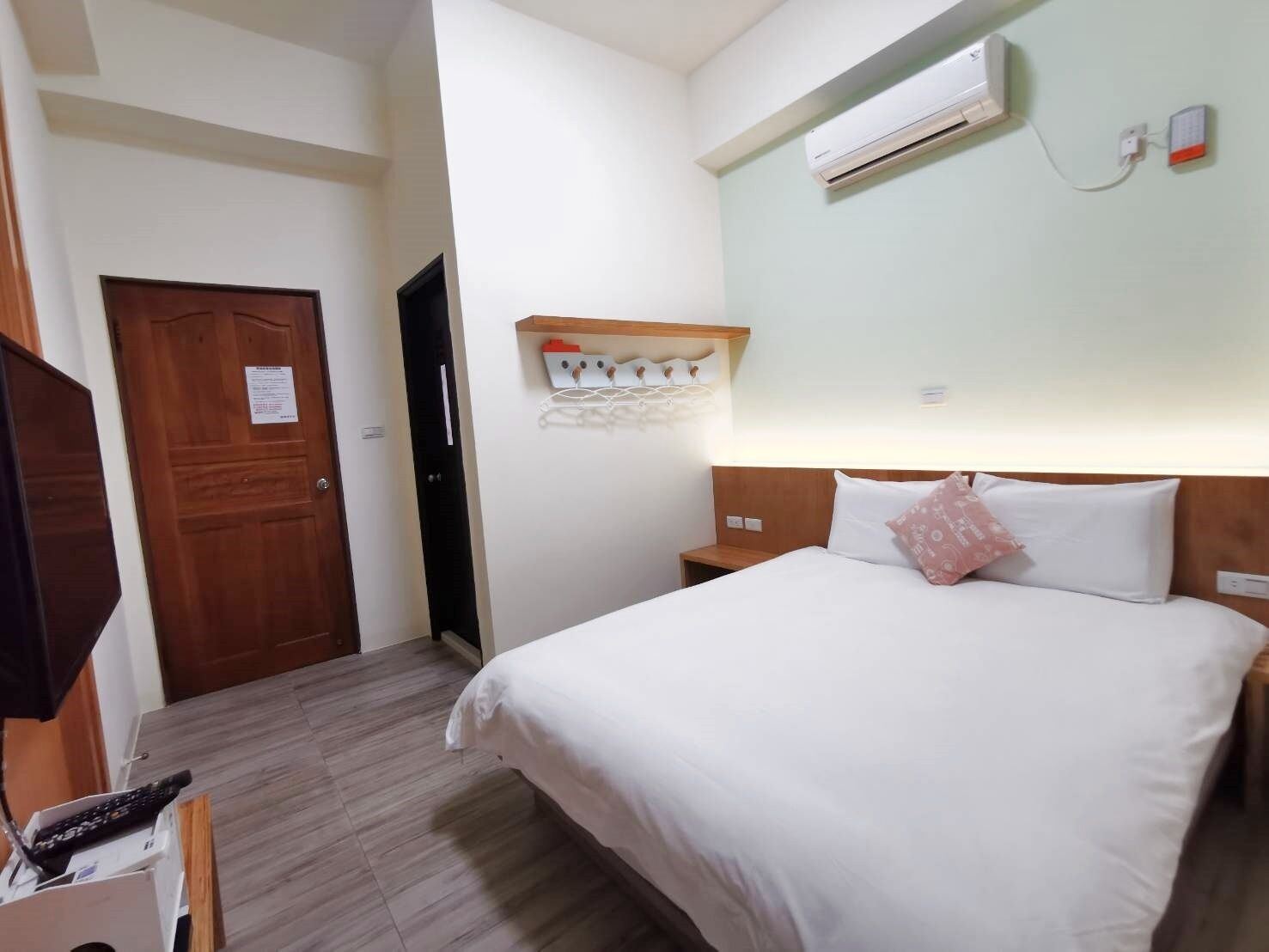 Una habitación de hotel con una cama, una puerta de madera, un aire acondicionado instalado en la pared y un pequeño estante con ganchos.