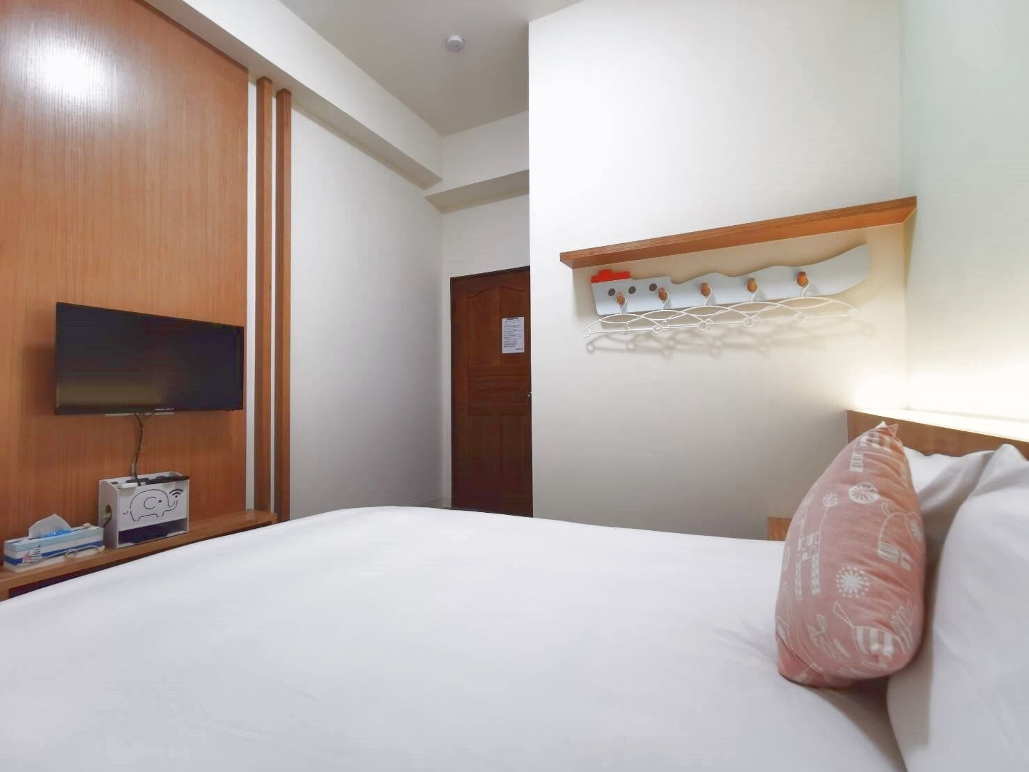 A hotel room with a bed, a television, a desk with a lamp, and a wall-mounted shelf with hooks.