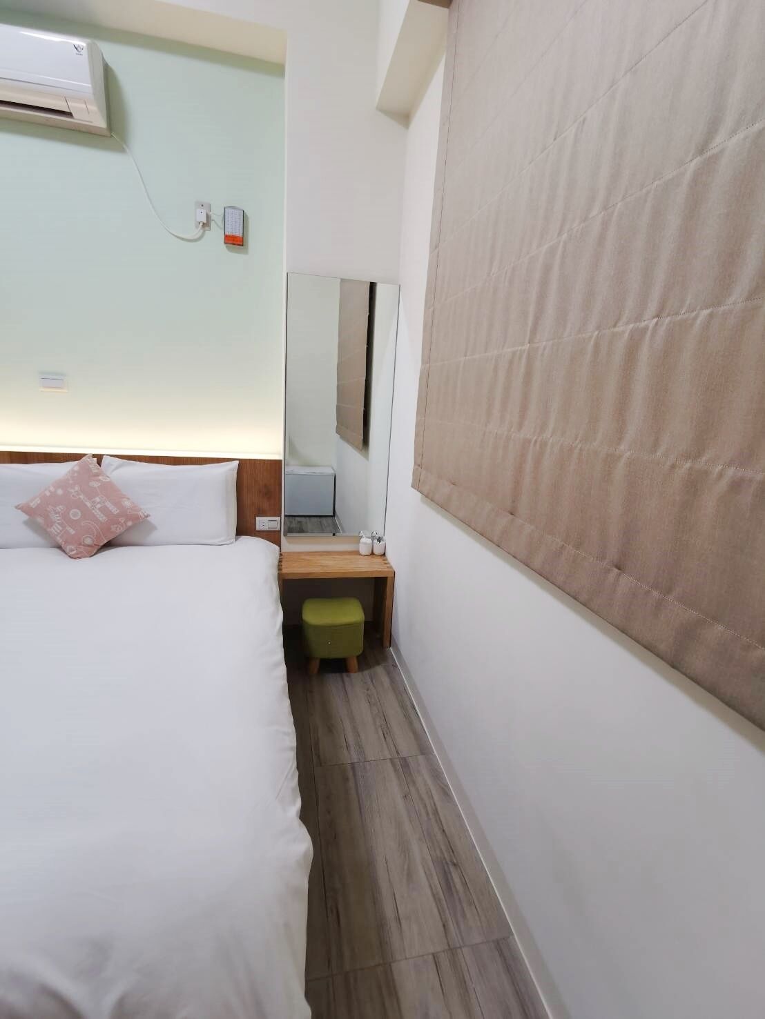 Una camera d'albergo con un letto, un comodino in legno, uno specchio e una finestra con una tenda beige.