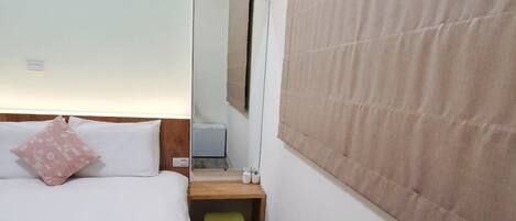 Una habitación de hotel con una cama, una mesita de noche de madera, un espejo y una ventana con persiana color beige.