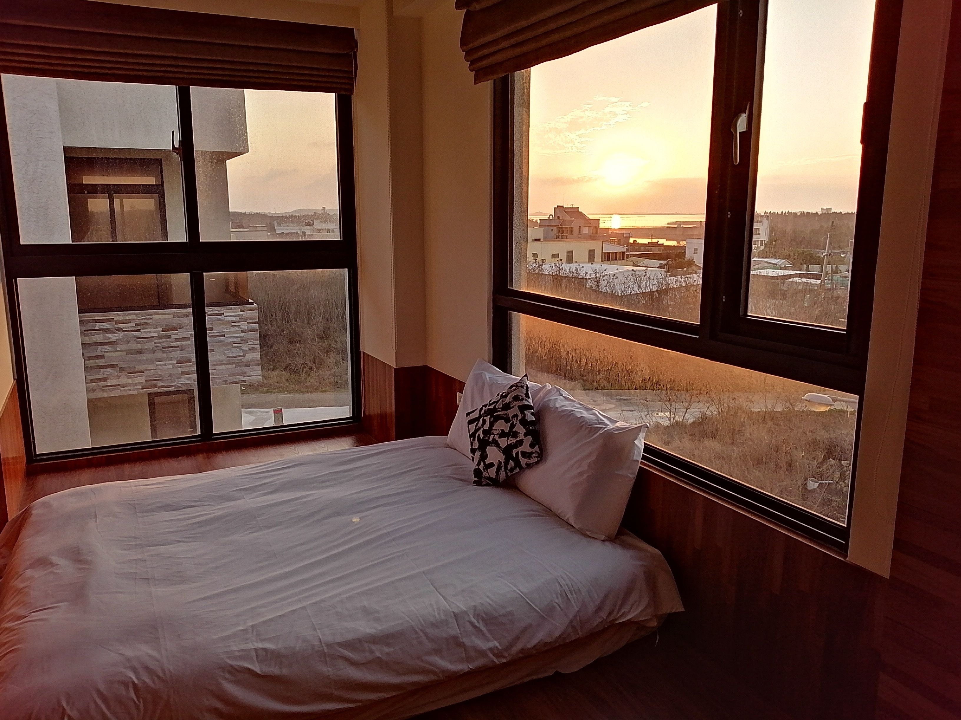 A bedroom with a large window offering a view of a cityscape and sunset.