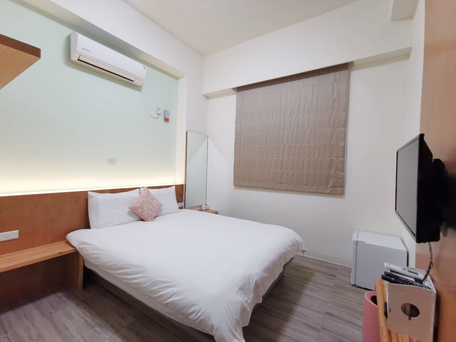 A hotel room with a bed, a bedside table, a TV, a window with blinds, and an air conditioning unit.