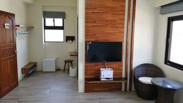 A modern living room with a flat-screen TV mounted on a wooden wall, a small stool, a wicker chair, and a glass table.