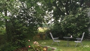 A garden with two chairs and a table, surrounded by lush greenery and flowering plants. - Vacation apartment Linde (2 persons) - Ferienwohnungen May (Plankenfels)