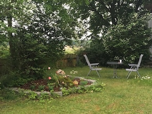 Un jardín con dos sillas y una mesa, rodeado de abundante follaje y plantas en flor.