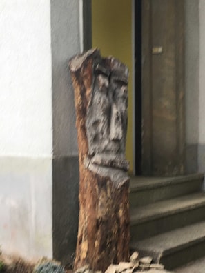 Una estatua de piedra de una figura con barba, ubicada sobre un pedestal frente a un edificio con puerta amarilla.