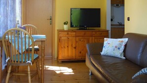 Uma sala de estar com piso de madeira, um sofá de couro marrom, uma mesa de jantar de madeira com cadeiras, uma televisão em um móvel de madeira e uma porta que dá para uma cozinha.