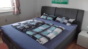 Quarto com cama, almofadas, mesa de cabeceira, abajur e janela com cortinas.