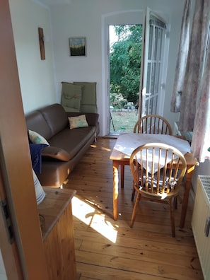 Un espacio acogedor con un sofá de cuero marrón, una mesa de comedor de madera y una silla. Se ve una ventana con cortinas, y una puerta da a un área exterior.