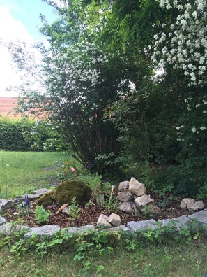 A garden with a variety of plants, including a tree with white flowers, a moss-covered rock, and a small wooden structure. - Vacation apartment Linde (2 persons) - Ferienwohnungen May (Plankenfels)