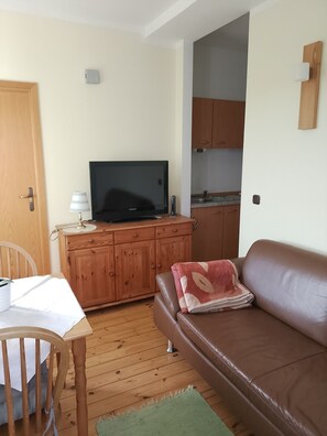 A cozy living room with a brown leather sofa, a wooden dining table with a chair, a television on a wooden cabinet, and a kitchen area visible through an open door. - Vacation apartment Linde (2 persons) - Ferienwohnungen May (Plankenfels)