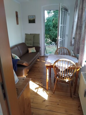 A cozy living area with a brown leather sofa, a wooden dining table, and a chair. A window with curtains is visible, and a door leads to an outdoor area. - Vacation apartment Linde (2 persons) - Ferienwohnungen May (Plankenfels)
