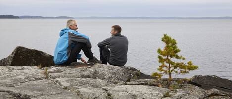 Dos personas sentadas sobre rocas a orillas de un lago, con un pequeño árbol cerca.