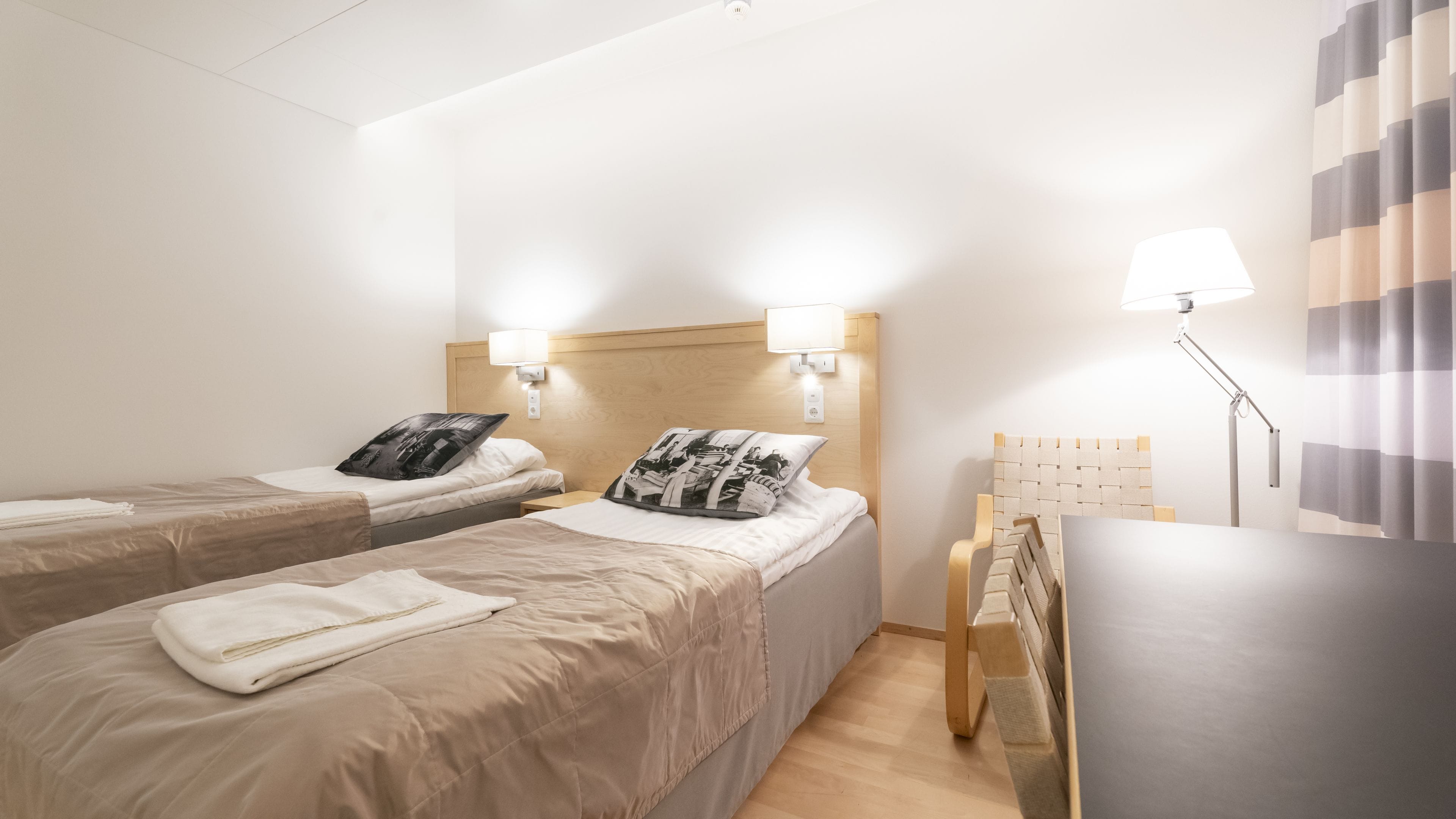 Een hotelkamer met twee bedden, elk voorzien van een kussen en een dekbed. Er is een bureau met een lamp en een stoel.