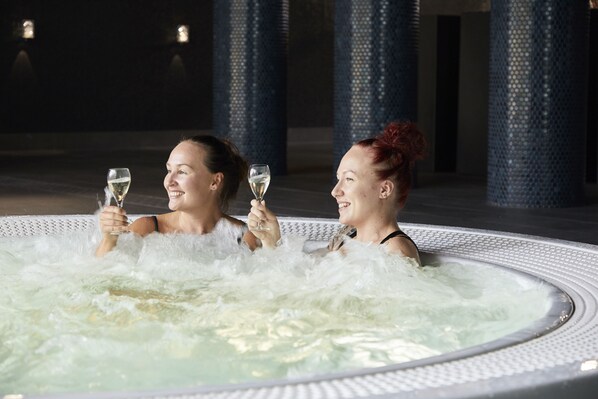 Dos personas en un jacuzzi con burbujas, sosteniendo vasos con un líquido transparente.