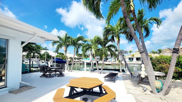 A patio area with a table and benches, surrounded by palm trees and a view of a marina with boats. - Fish Heaven- 2 bed, 2 bath in KCB with boat lift, ice machine & cabana club (Key Colony Beach)