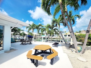 A patio area with a table and benches, surrounded by palm trees and a view of a marina with boats. - Fish Heaven- 2 bed, 2 bath in KCB with boat lift, ice machine & cabana club (Key Colony Beach)