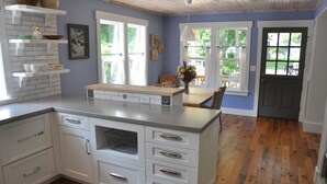 Une cuisine dotée de meubles blancs, d’un plan de travail en bois et offrant une vue sur l’extérieur grâce à de grandes fenêtres.