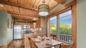 Una cocina de madera con una mesa de comedor para cuatro personas, un refrigerador y un ventanal con vista a árboles.
