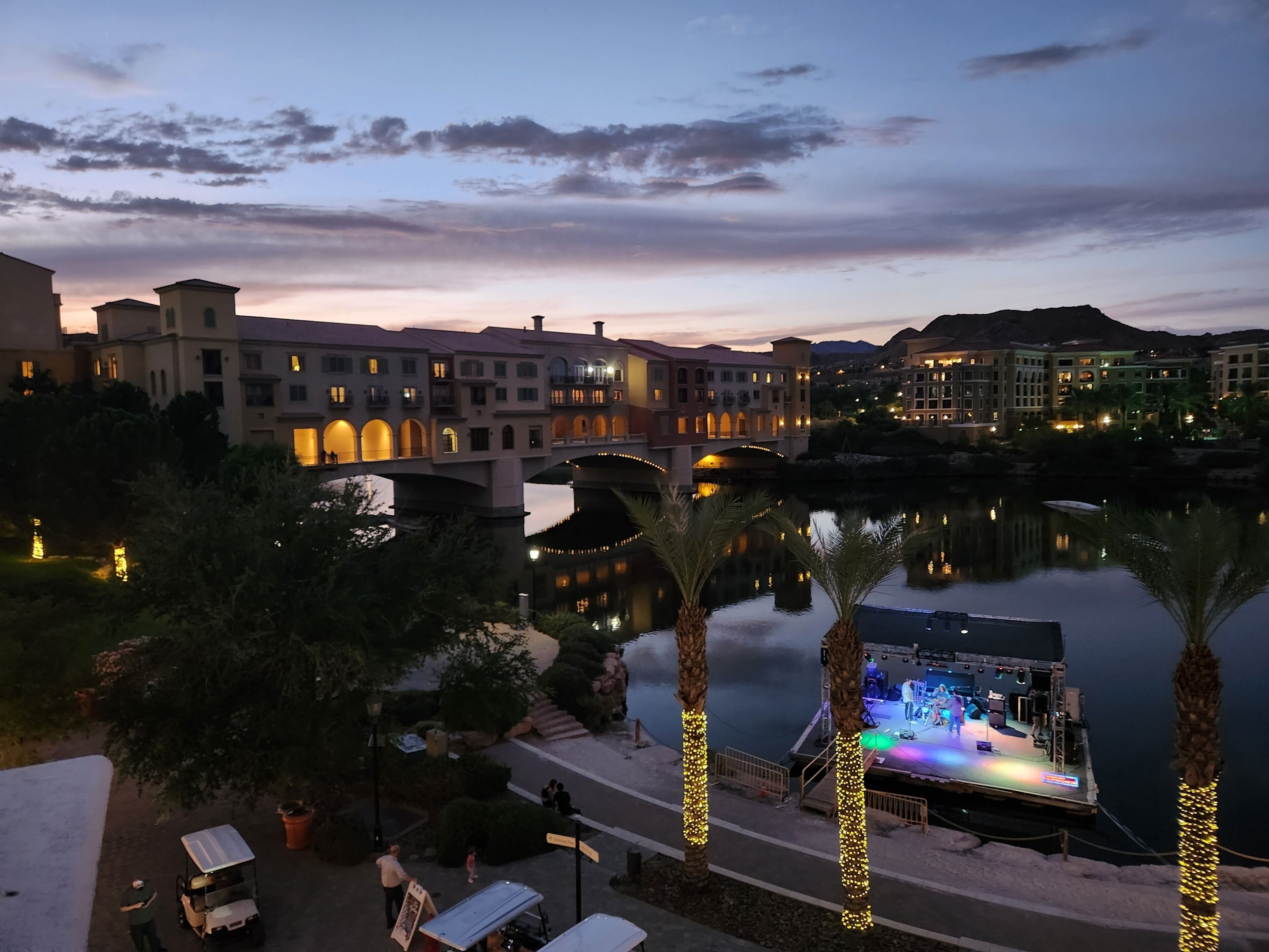 A riverside hotel with a stage for events, palm trees, and a bridge.