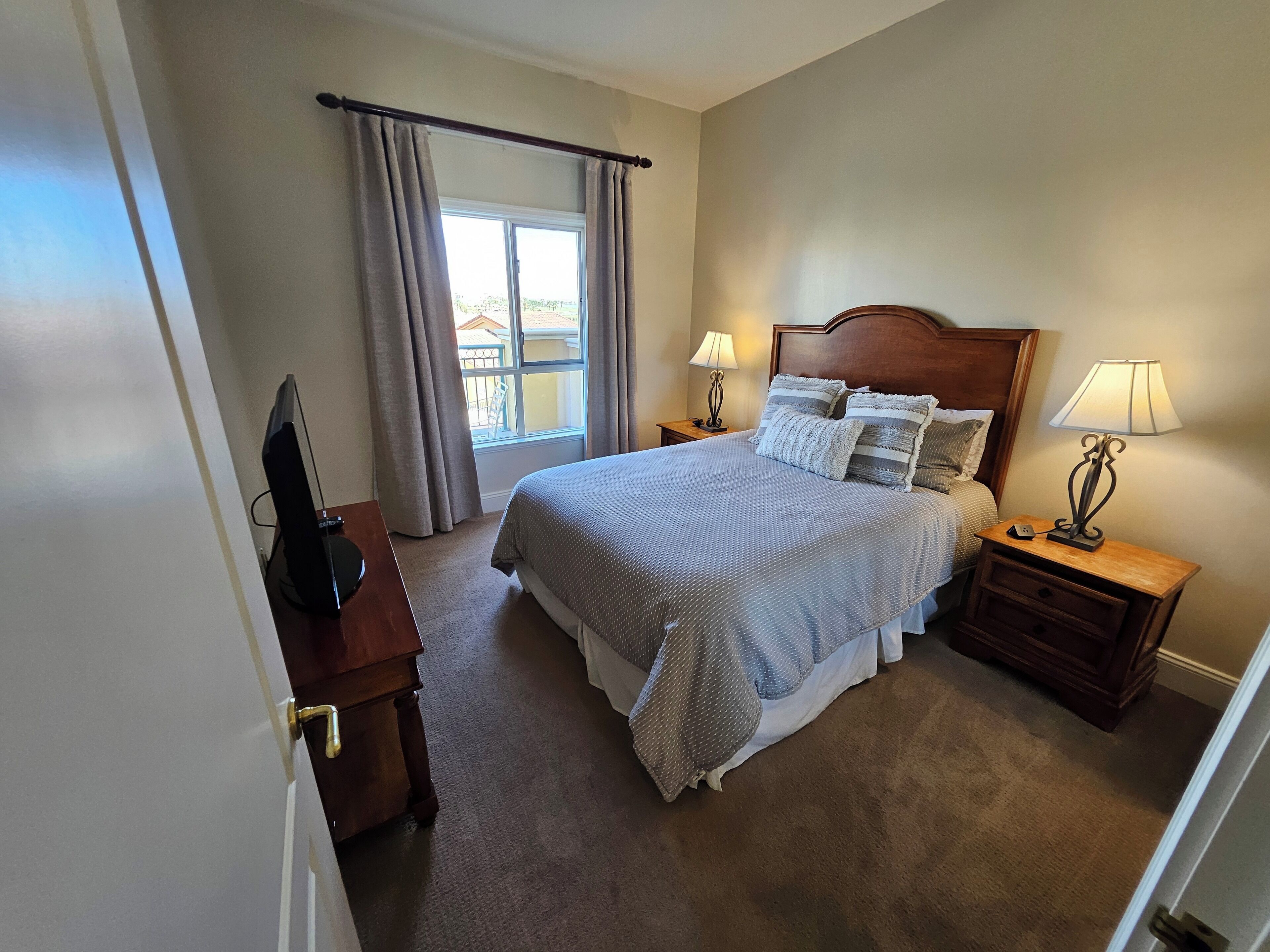 A neatly arranged bedroom with a large bed, two bedside tables with lamps, a window with curtains, and a television on a wooden stand.