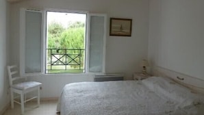 A bedroom with a bed, a chair, a window with a view of greenery, and a framed picture on the wall.