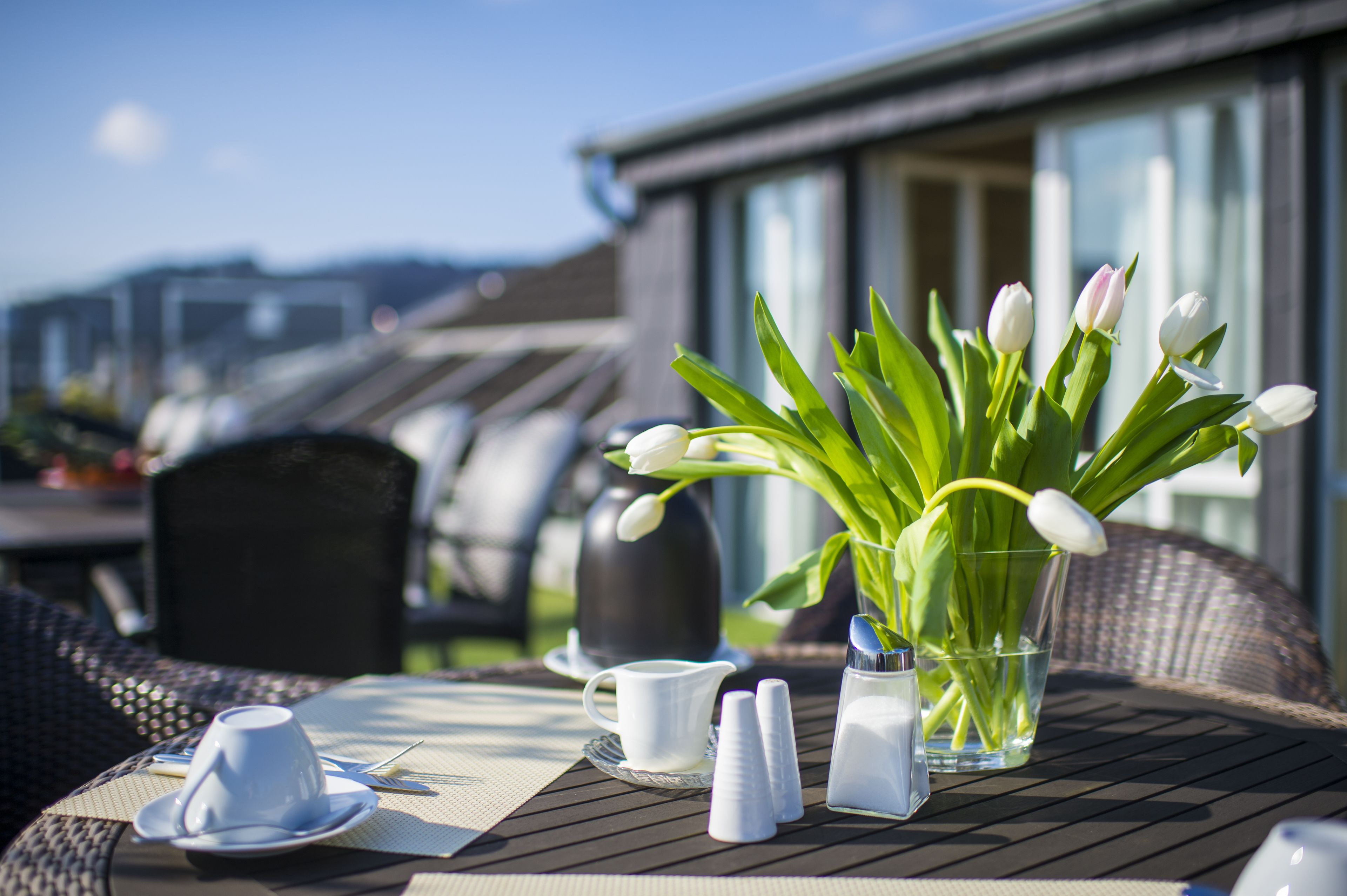 Ein Tisch ist gedeckt mit weißen Tassen, einer Kanne, Salzstreuern und einer Vase mit weißen Tulpen auf einer sonnigen Terrasse mit Korbmöbeln und Blick auf ein Gebäude.