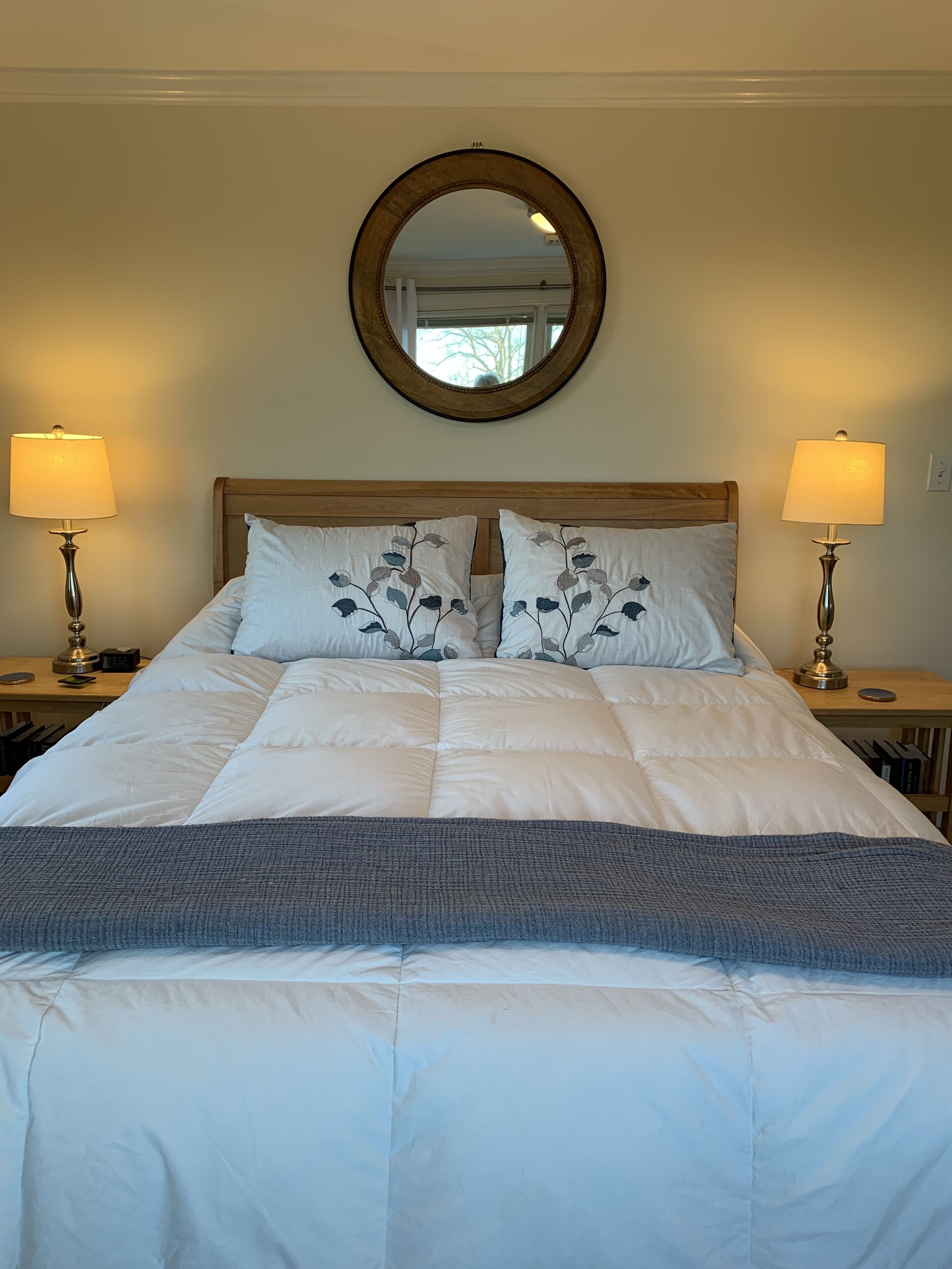 A neatly made bed with a wooden headboard, two bedside lamps, and a round mirror above.