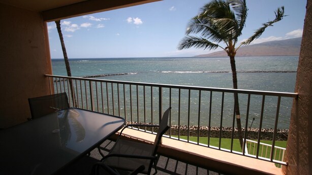 Un balcon avec une table et des chaises en verre, donnant sur une plage bordée de palmiers et sur l’océan.