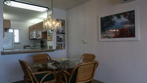 Un coin repas avec une table en verre, des chaises en osier, un lustre, une cuisine en arrière-plan et un tableau encadré au mur.